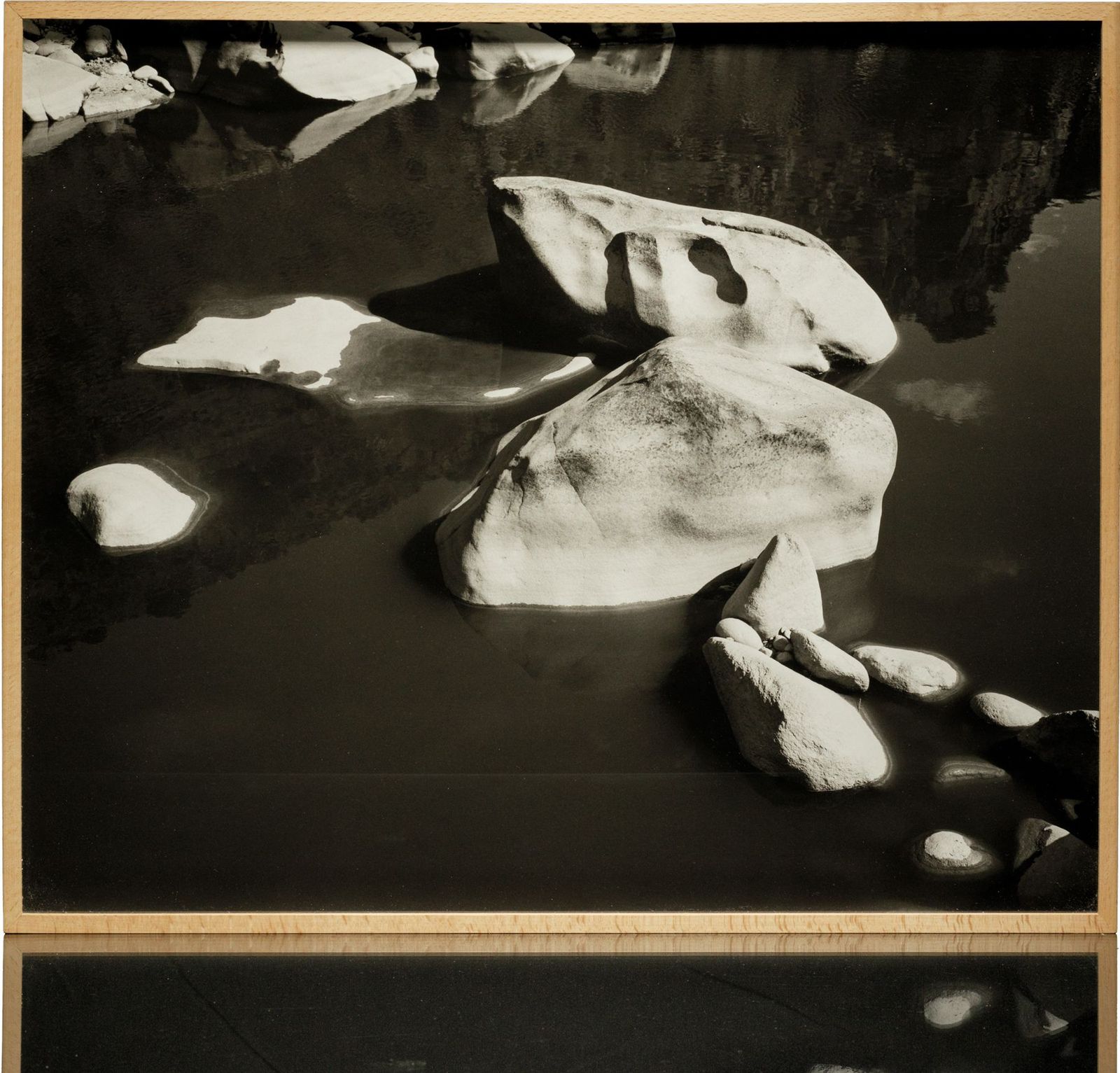 A framed black-and-white print: pale boulders in dark, still water, in a light wood frame