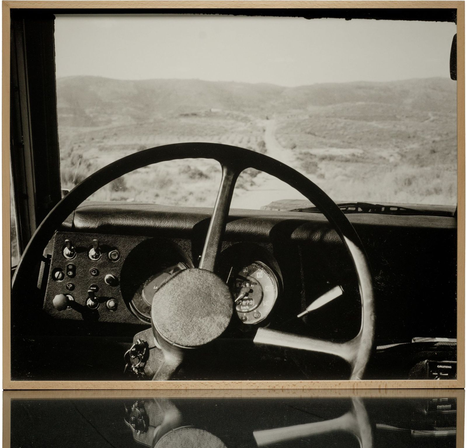 A framed black-and-white print: the steering wheel and dashboard of an old vehicle, a dirt road running into the hills beyond the windscreen, in a light wood frame