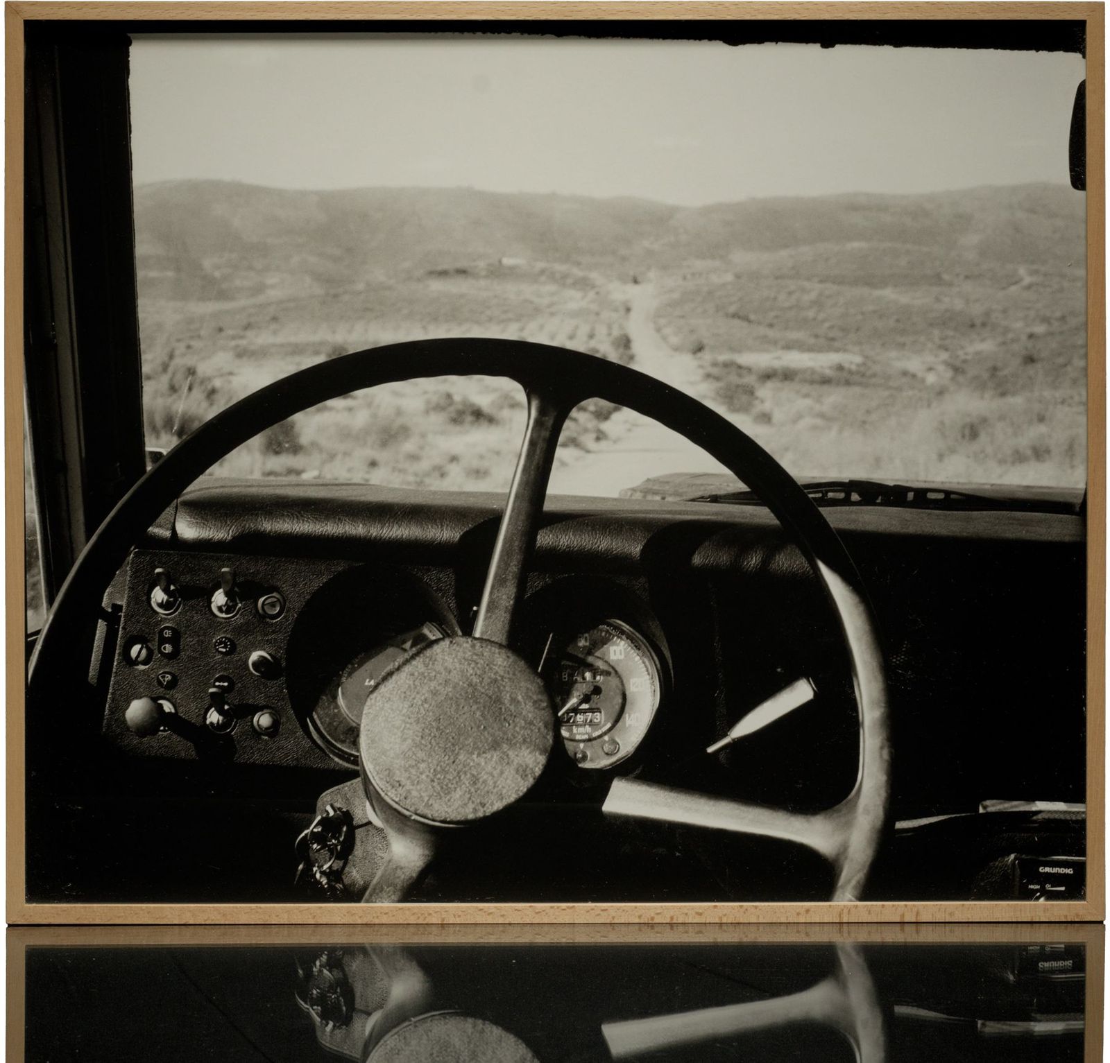 A framed black-and-white print: the steering wheel and dashboard of an old vehicle, a dirt road running into the hills beyond the windscreen, in a light wood frame