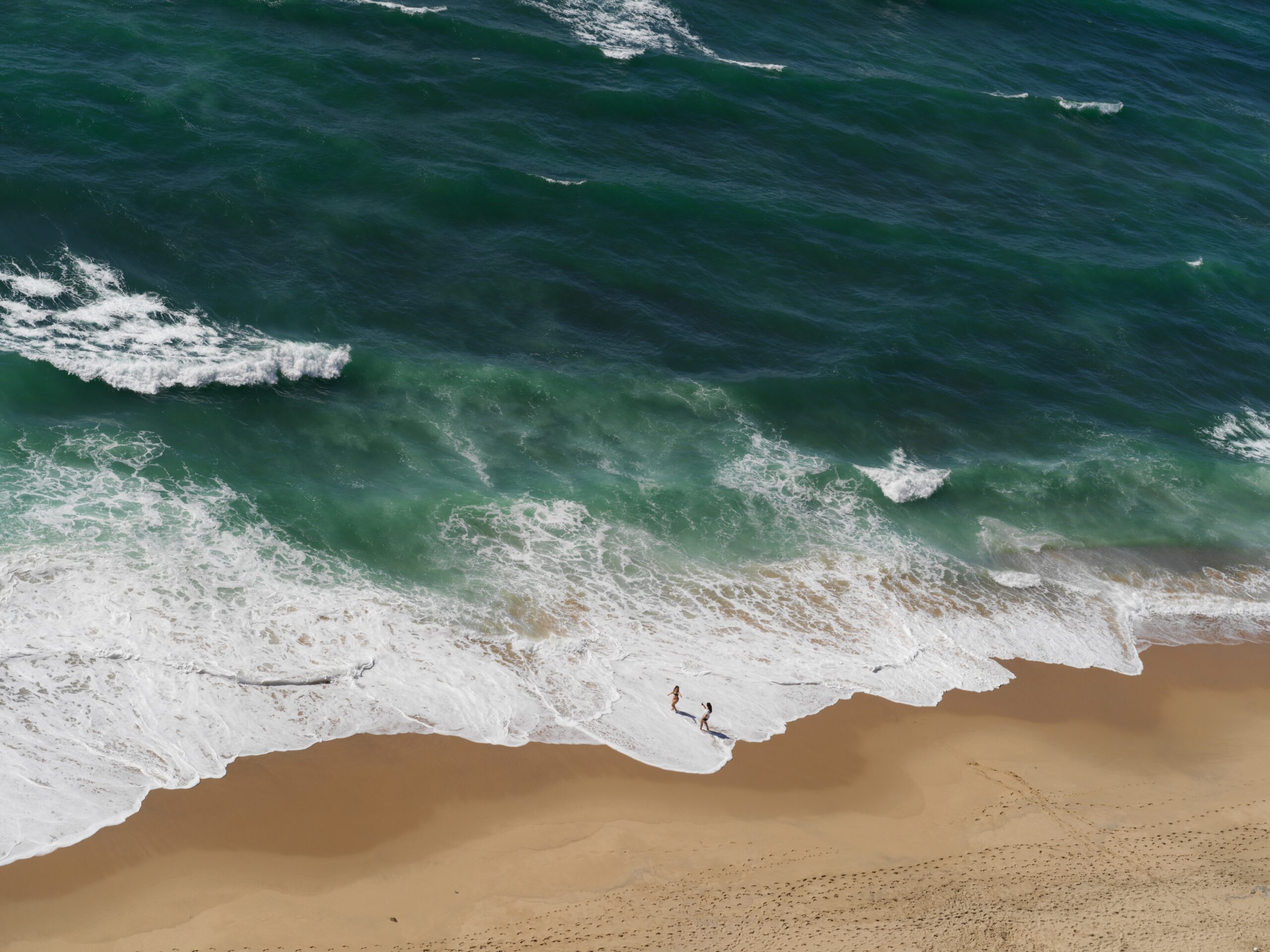 Rebentação numa praia, vista de cima, com duas figuras na orla — fotografia de Pedro Guimarães